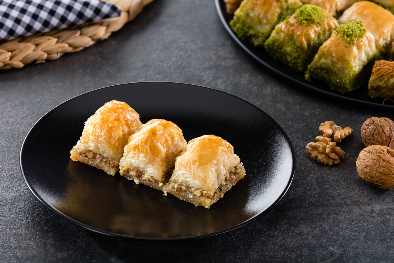 Three baklava pieces on a black plate with walnuts on a dark surface.